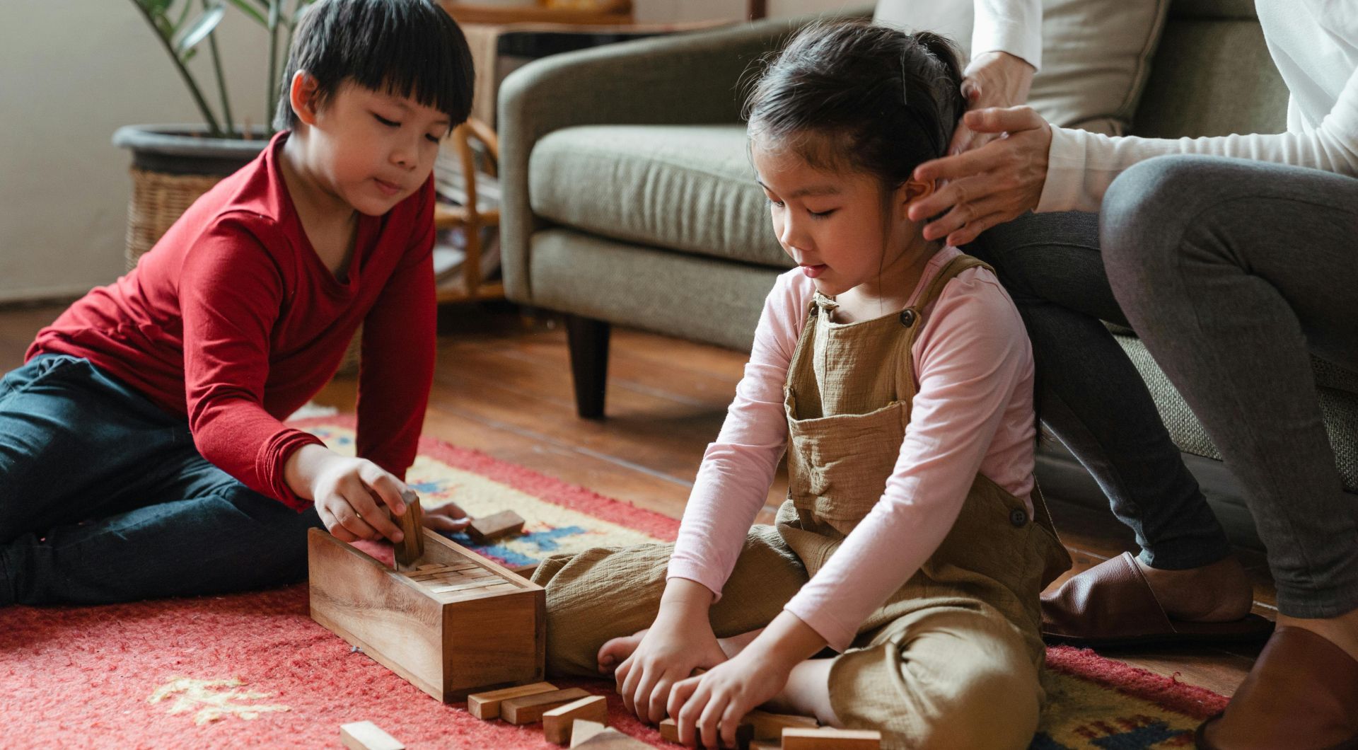 Unrecognizable mother sitting on sofa in modern living room while spending time together with Asian son and daughter sitting on floor carpet and playing jenga game